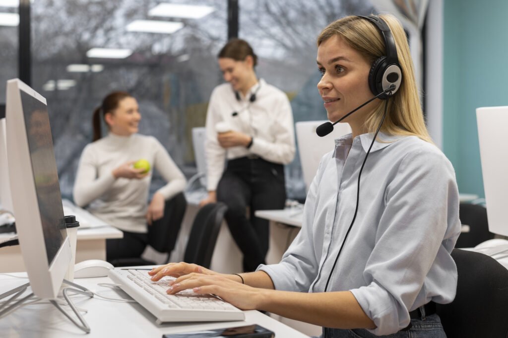 A blonde girl wearing her headset and typing in front of her laptop and two employees chatting in the background