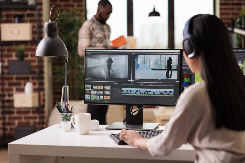 A woman wearing her headset working on her montaging software editing creative footage and an employee standing in the background wearing casual