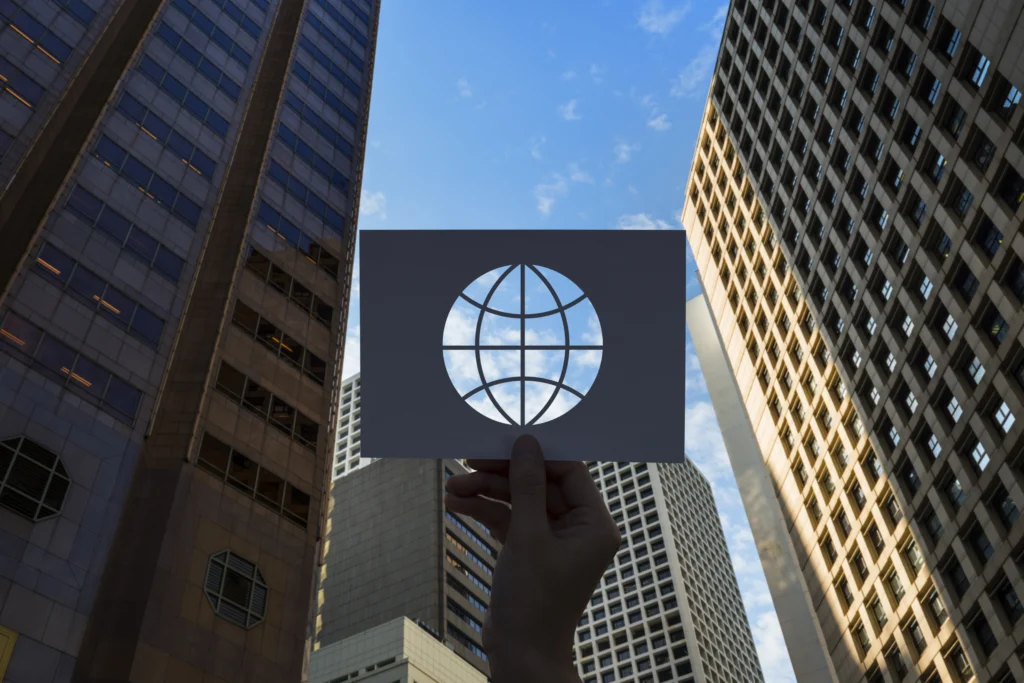 A wide-angle image of a person holding a paper cutout in the shape of the world, with tall buildings and a vibrant sky in the background. The scene emphasizes a blend of creativity, global vision, and modern urban life