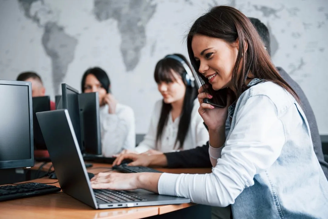 A woman happily answering calls in her customer support job, with engaged employees working in the background.