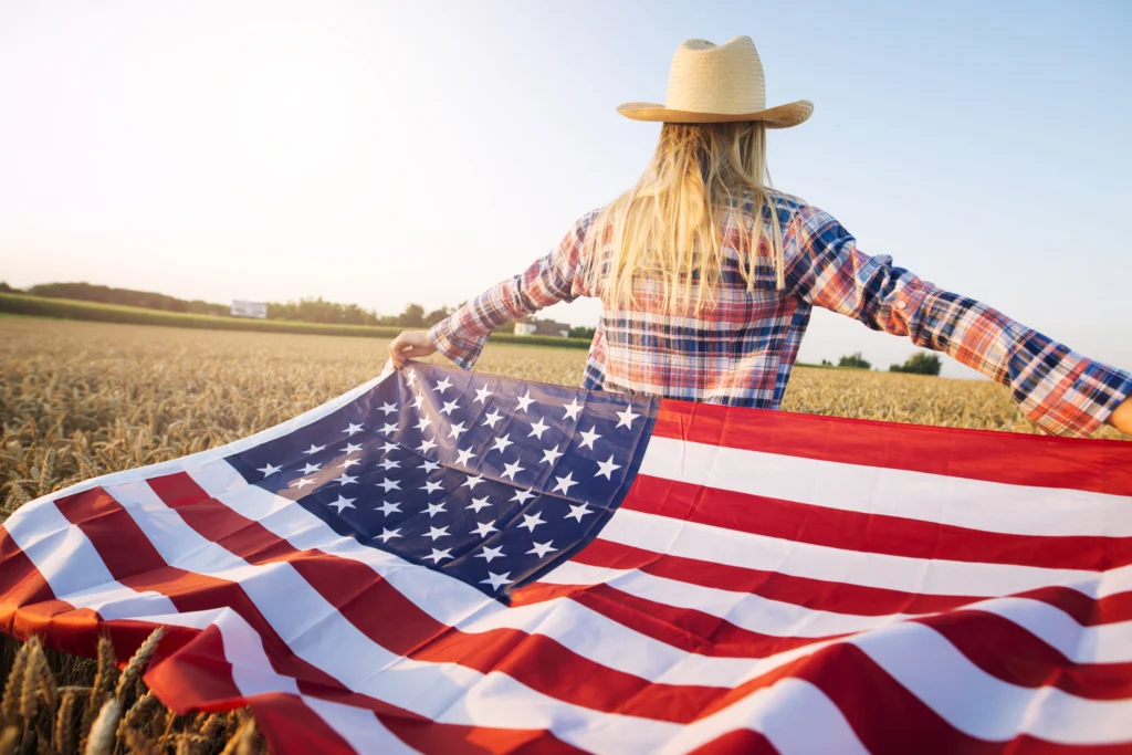 A female farmer stands in a wheat field, her back turned to the camera, with her arms spread open holding an American flag. She wears a plaid shirt and a straw hat, with golden sunlight illuminating the scene. The expansive field stretches into the horizon under a clear sky, symbolizing rural American pride and freedom.