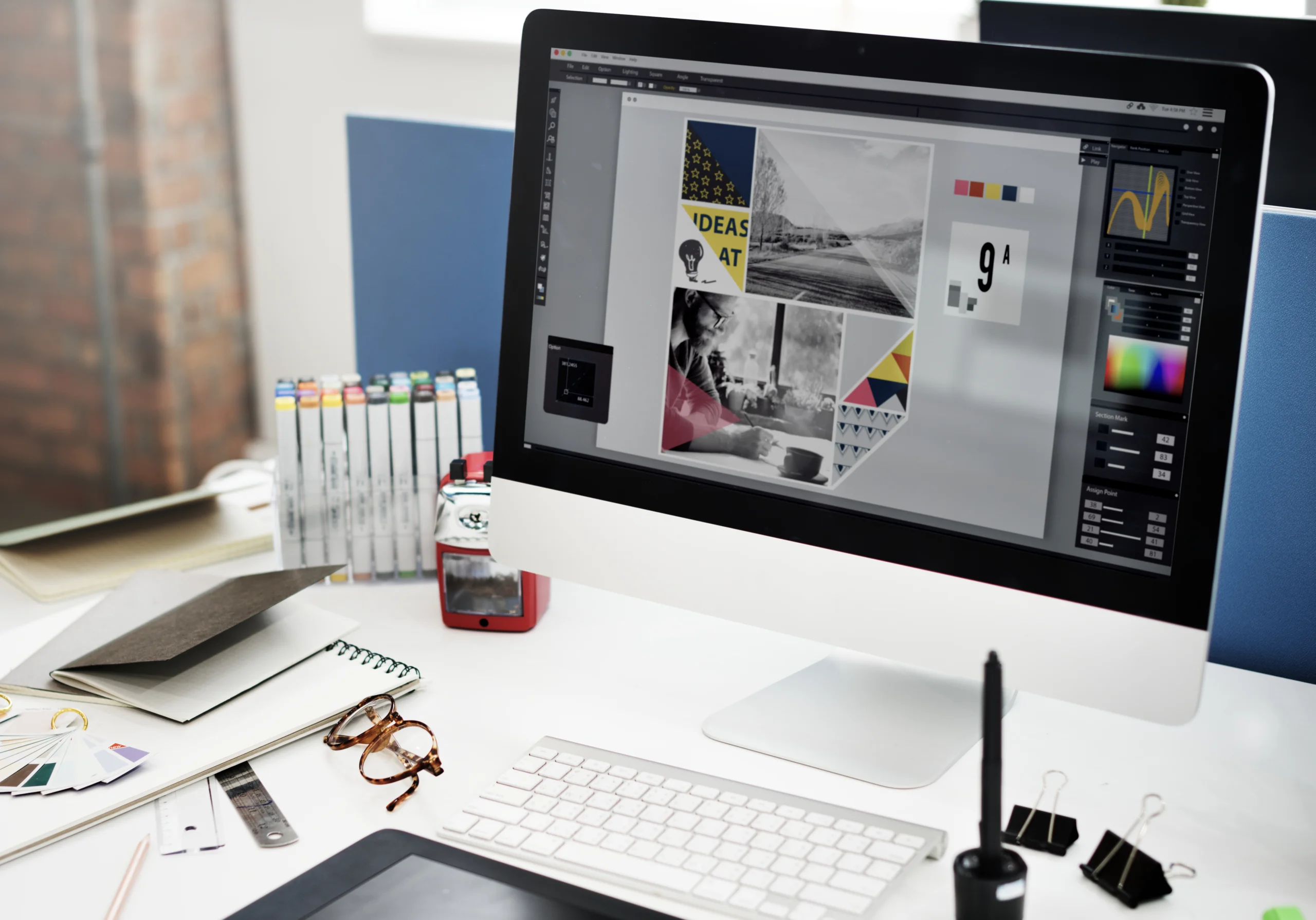 A sleek white-themed PC setup features a monitor displaying software filled with logos, design layouts, and various documents. The desk is organized with glasses, stationery, and other work essentials, creating a clean and productive workspace that showcases creativity and professionalism.