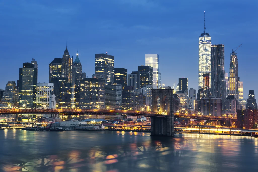 A cinematic sunset view of Manhattan, New York, as the city's skyscrapers and buildings start to light up, casting a warm glow over the skyline. The iconic bridge stands prominently in the foreground, bathed in soft evening light, with a vivid array of illuminated windows and streetlights beginning to shine.