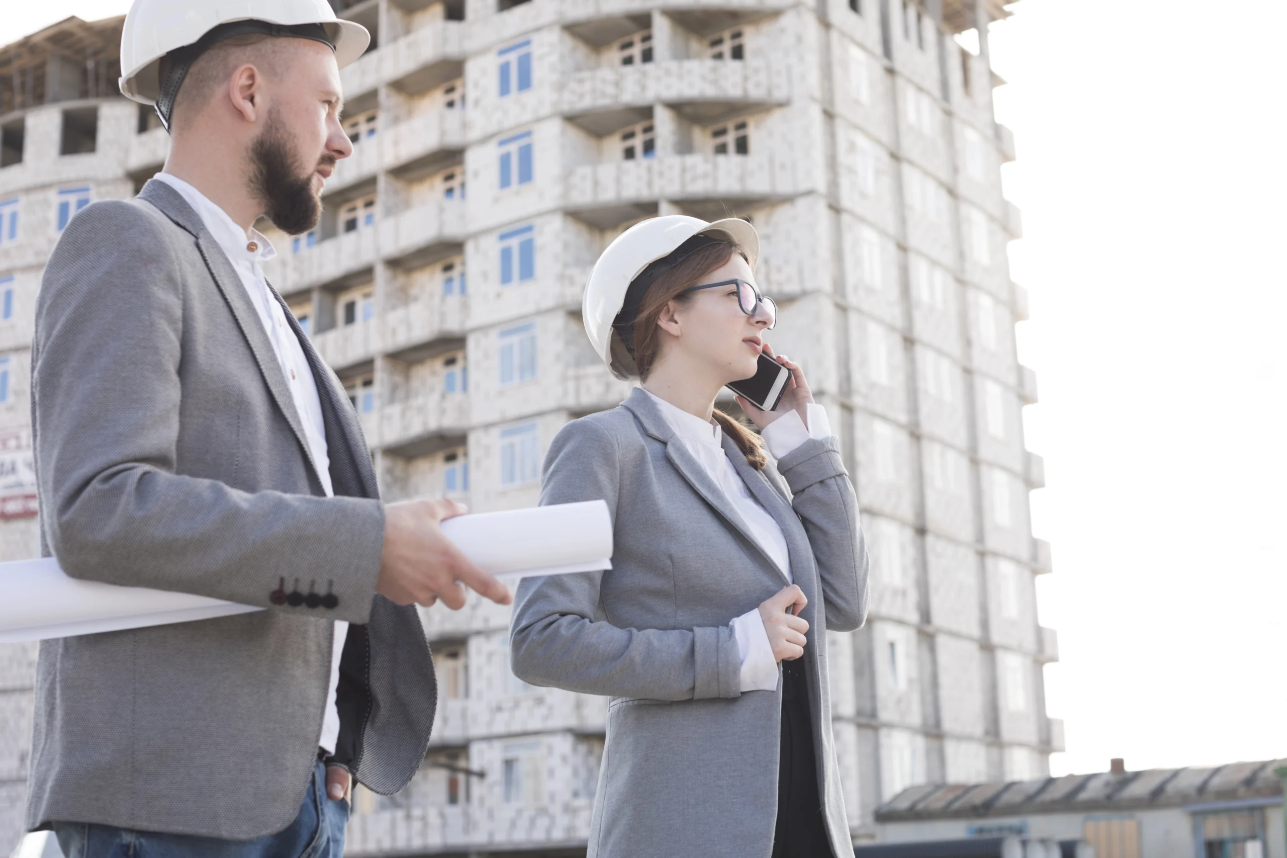 A business engineer holds a blueprint while a female business engineer speaks on her phone. Both are dressed in matching suits, showcasing professionalism, with construction buildings rising in the background, symbolizing collaboration and progress in their projects.






