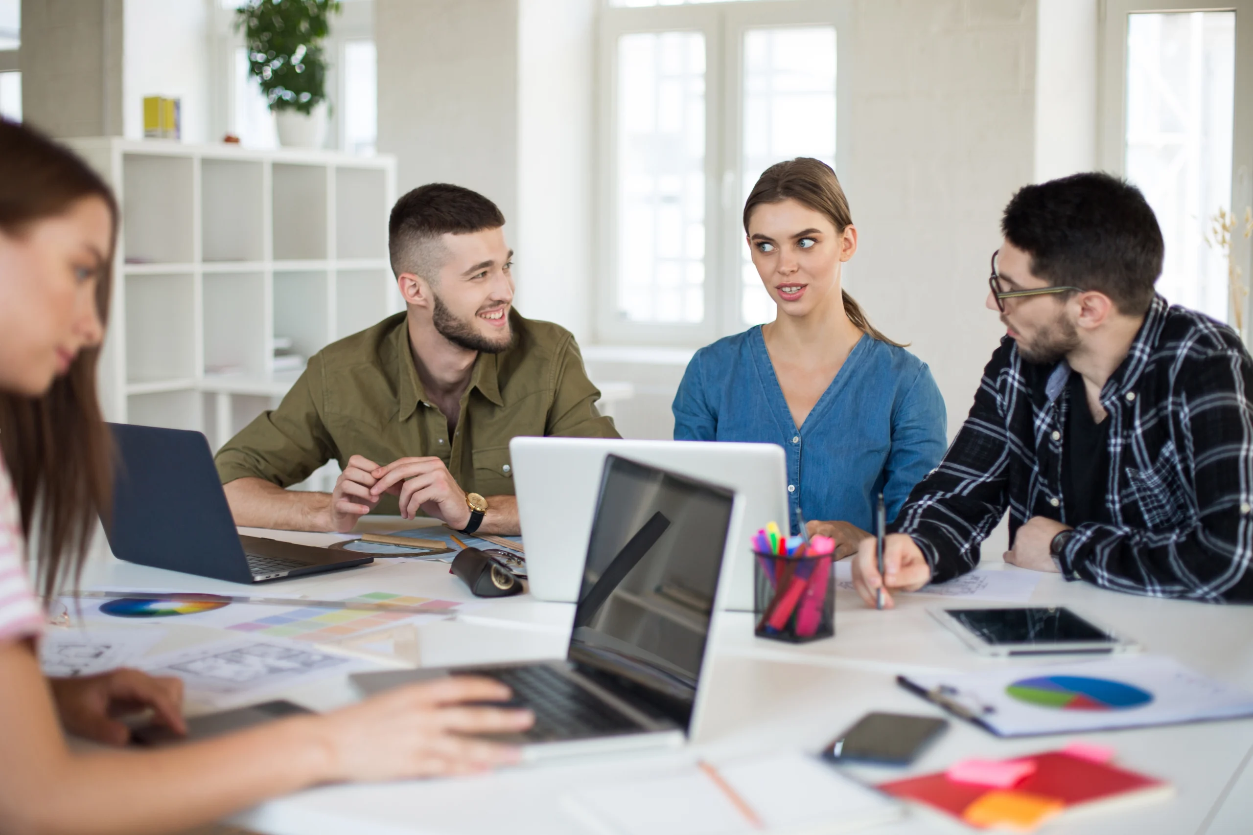 Four employees at a web design agency working together around a meeting table, using laptops, papers, and prototypes for project development.